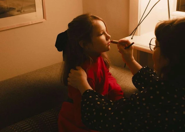Young child in a red top having lipstick applied by an adult in a polka-dot dress inside a dimly lit room