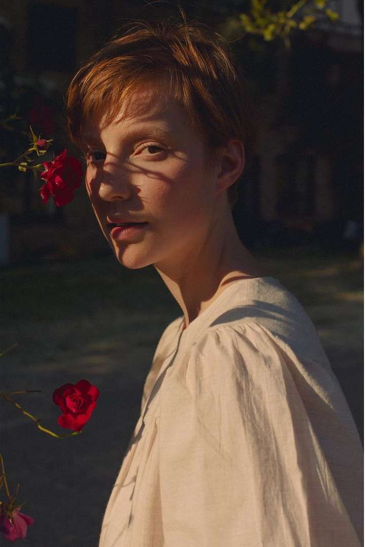 Close-up portrait of a fair-skinned woman with short red hair, standing outdoors next to red roses in diffused sunlight.