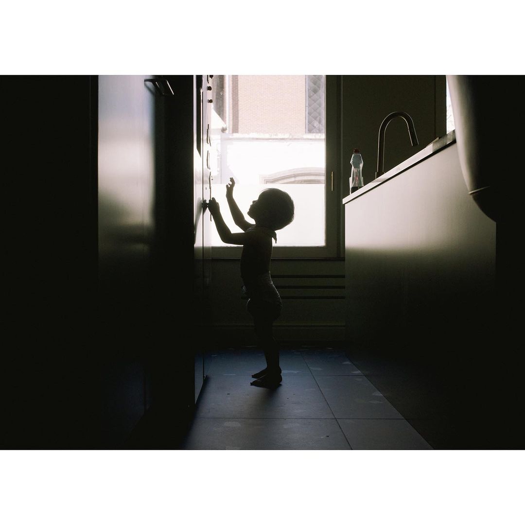 Silhouette of a toddler reaching up in an interior with natural light streaming in from a window, adjacent to kitchen cabinets.