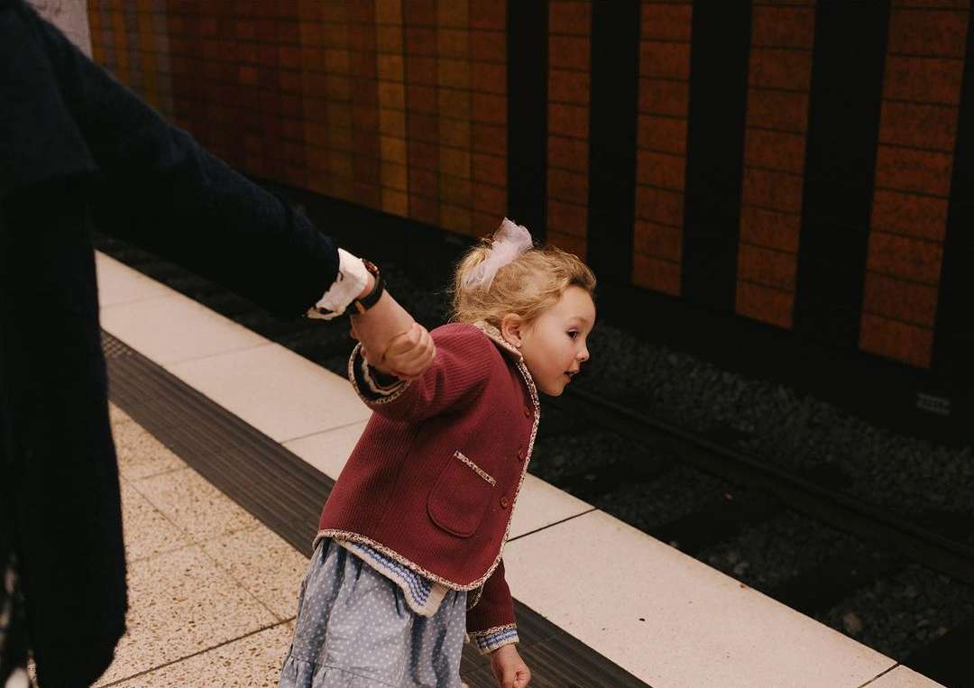 Little girl in red jacket holds adult's hand on train platform, looking at subway tracks with yellow tiled background.