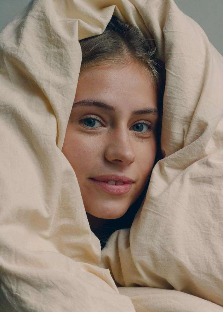 A young woman with blue eyes and light brown hair is enveloped in a cream-colored blanket, looking directly at the camera.