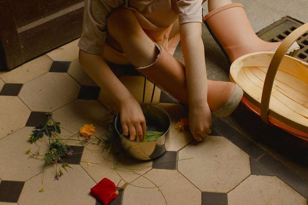 A young child sits on a tiled floor, wearing pink boots, playing with leaves and flowers in a metal pot, next to a wooden basket.