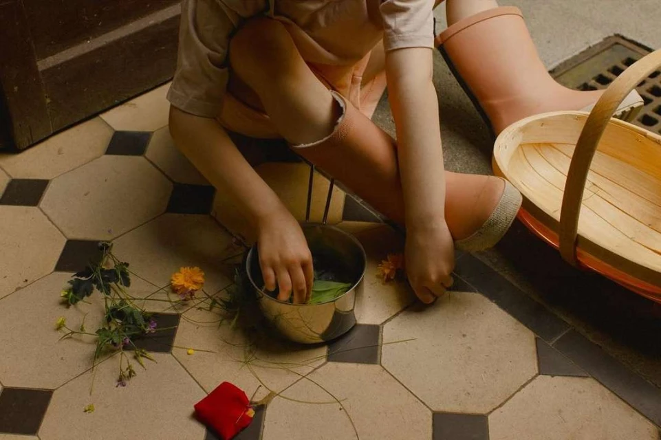 A young child sits on a tiled floor, wearing pink boots, playing with leaves and flowers in a metal pot, next to a wooden basket.