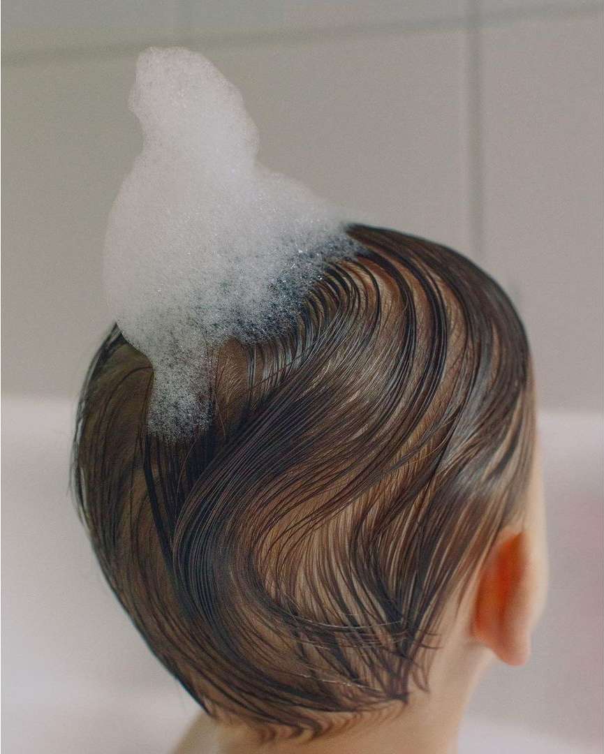 Close-up of a baby's head with brown hair covered in white soap bubbles in a bright bathroom setting, showcasing childhood bath time.