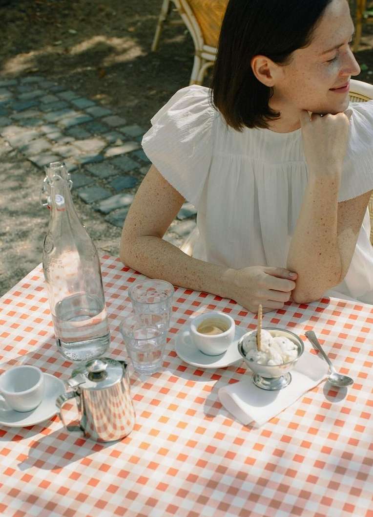 Woman in white dress sits at outdoor cafe table with espresso, water, and ice cream on a red-and-white checkered tablecloth.