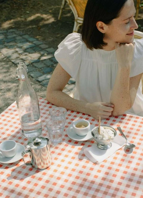 Woman in white dress sits at outdoor cafe table with espresso, water, and ice cream on a red-and-white checkered tablecloth.