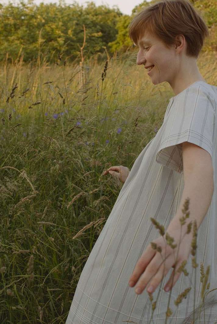 Woman in a striped dress walks through a field of tall grass on a sunny day, smiling slightly and extending her arms.