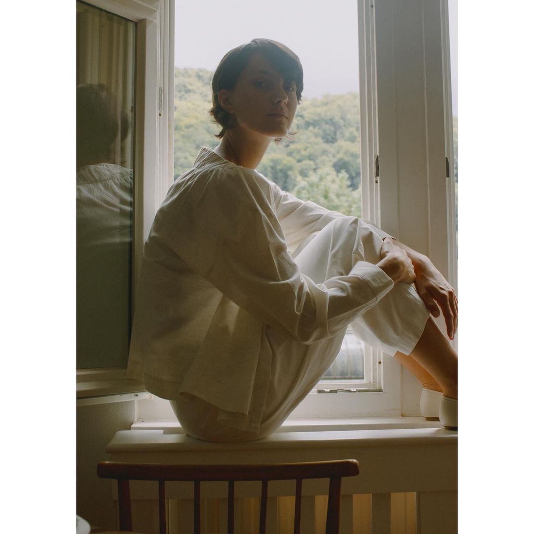 Adult female in white sitting in a window wearing matching shirt and pants with rural scenery visible in background during the daytime.
