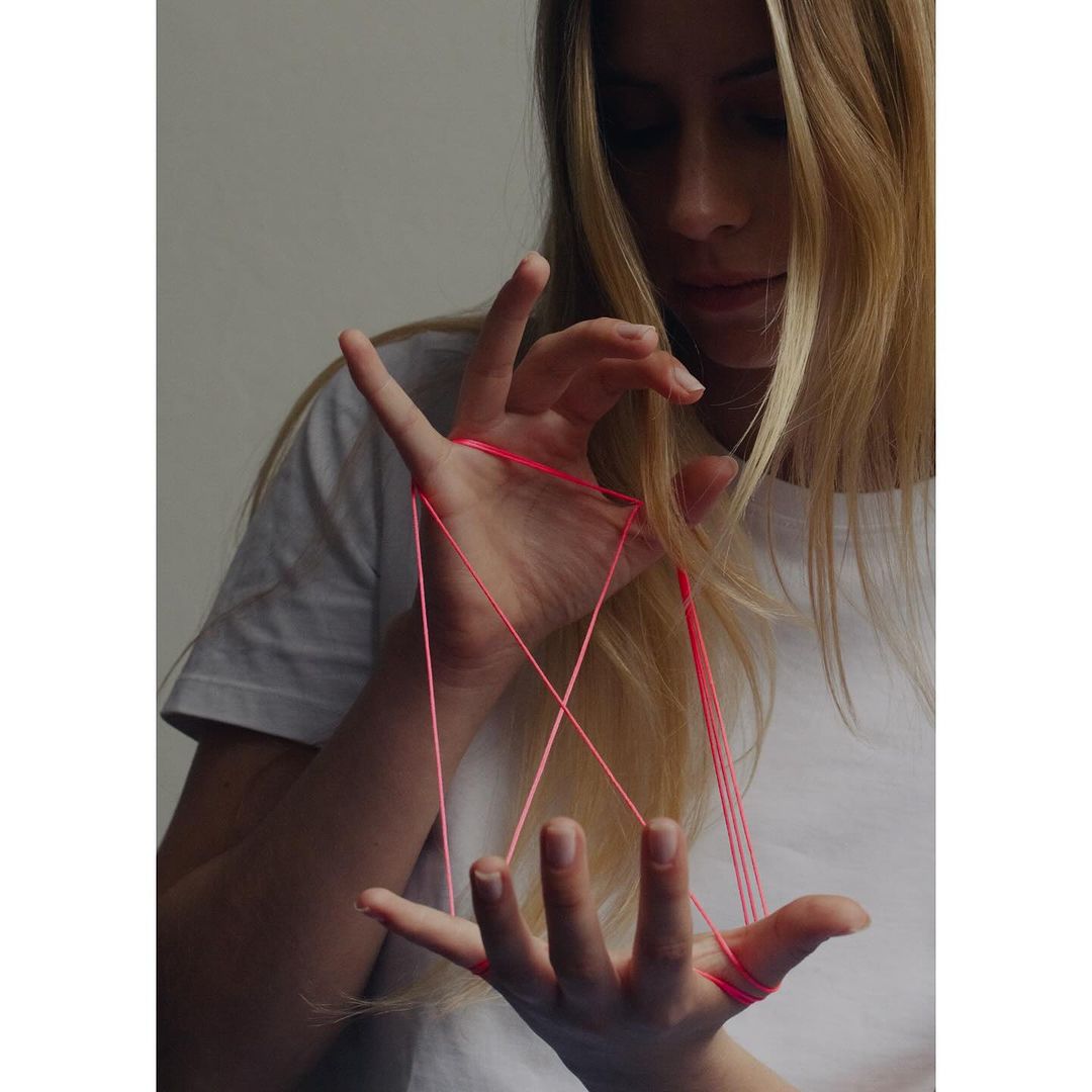 Young woman with long blonde hair plays cats cradle with red string. The indoor scene is softly lit.