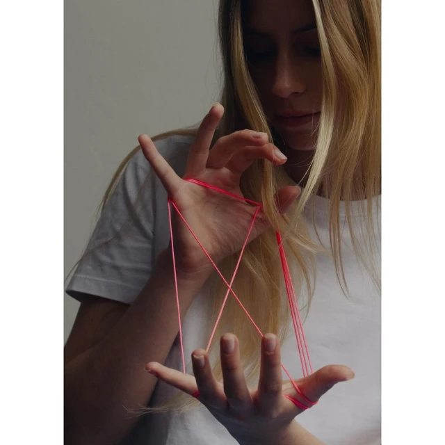 Young woman with long blonde hair plays cats cradle with red string. The indoor scene is softly lit.