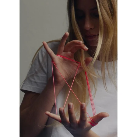 Young woman with long blonde hair plays cats cradle with red string. The indoor scene is softly lit.