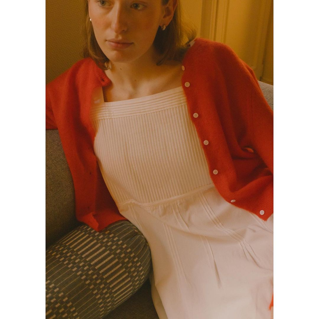 Young adult woman wearing a red cardigan and white dress indoors, seated on a gray couch. Warm lighting adds to the cozy atmosphere.