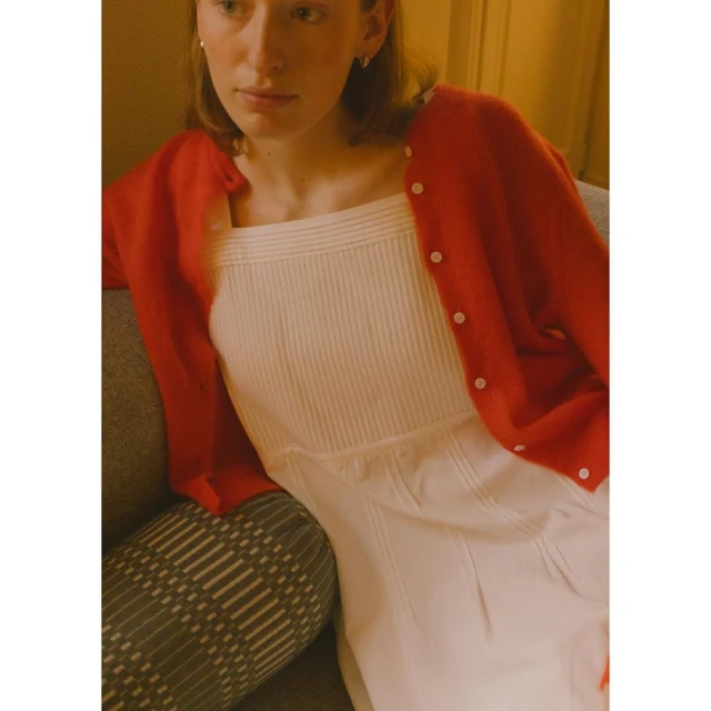 Young adult woman wearing a red cardigan and white dress indoors, seated on a gray couch. Warm lighting adds to the cozy atmosphere.