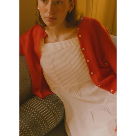 Young adult woman wearing a red cardigan and white dress indoors, seated on a gray couch. Warm lighting adds to the cozy atmosphere.