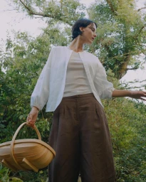 Woman in white shirt and brown trousers holding a wooden basket outdoors in a leafy garden.