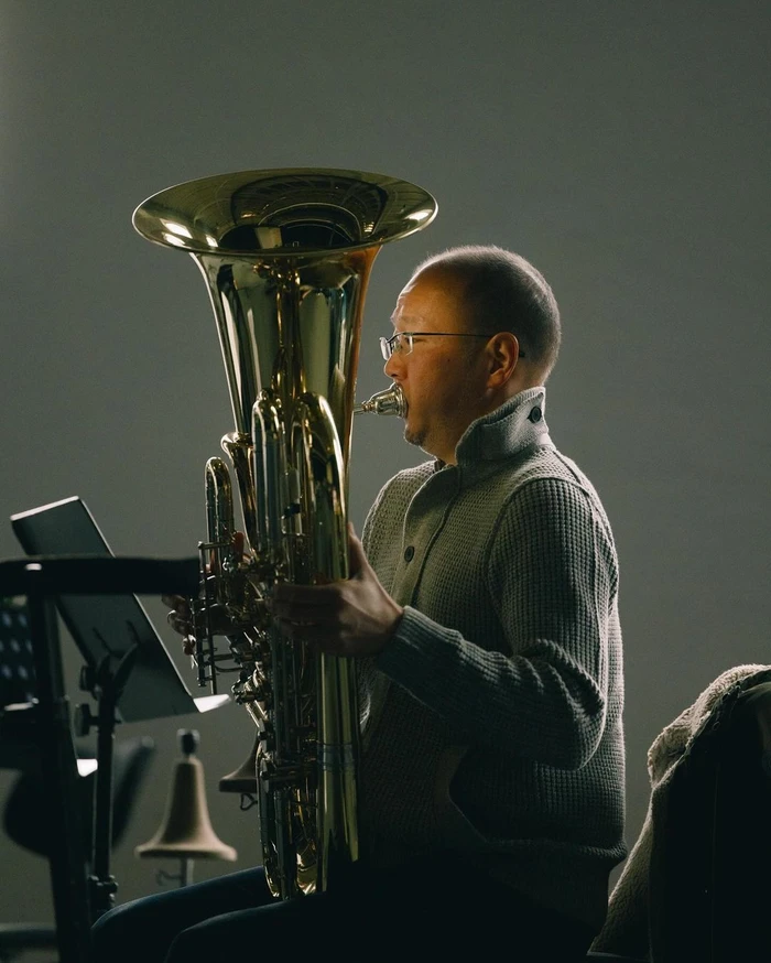 Man in grey sweater wearing glasses playing a large brass tuba indoors