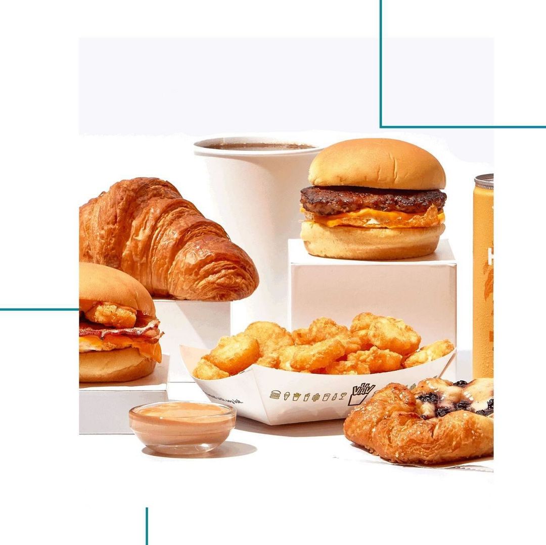 Still life featuring a croissant, burgers, hash browns, a white coffee cup, danish pastry, and a drink can, on a white surface.