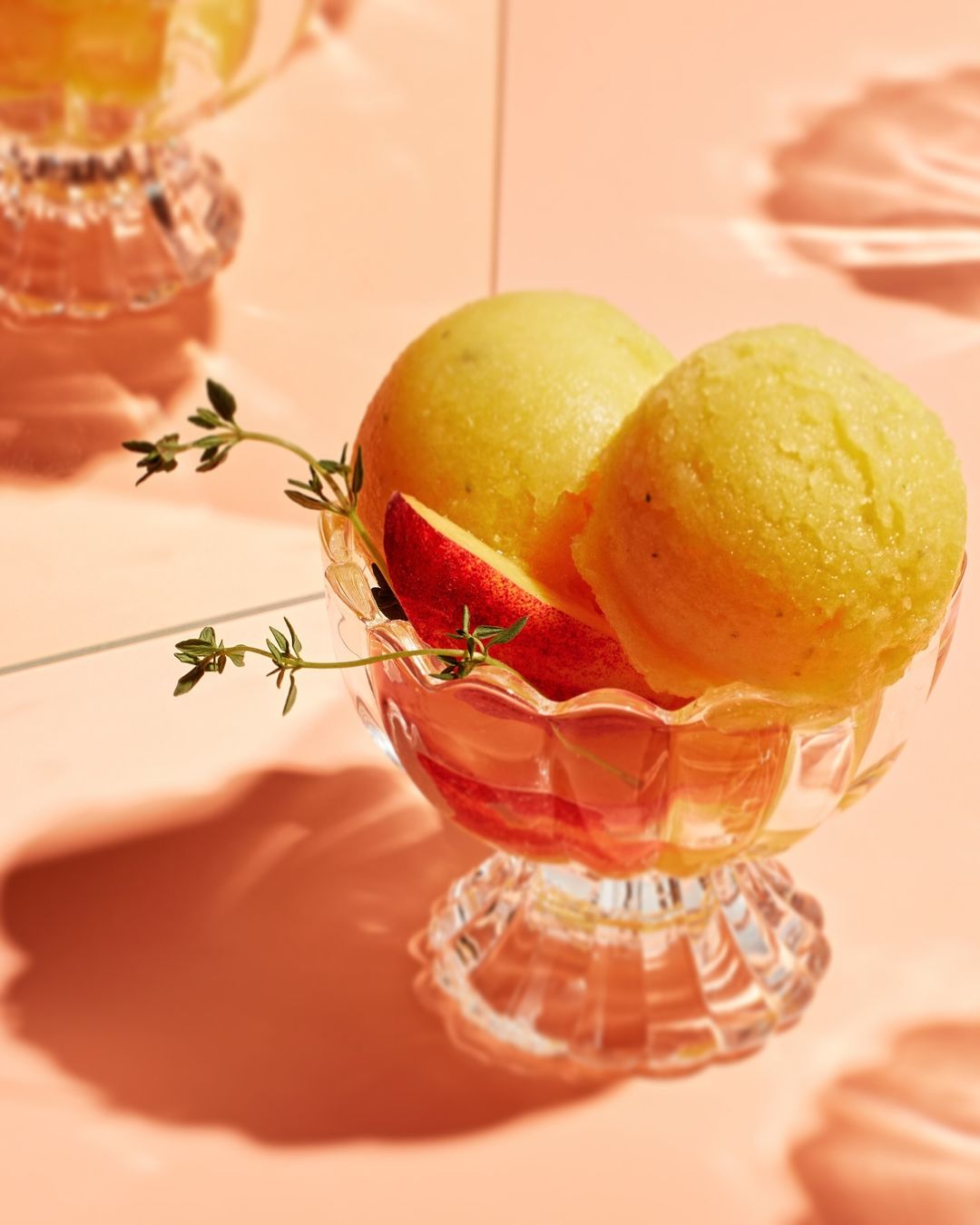 Close-up of yellow sorbet scoops served in a clear glass dessert cup with peach slice and thyme sprigs, against a peach-colored background.