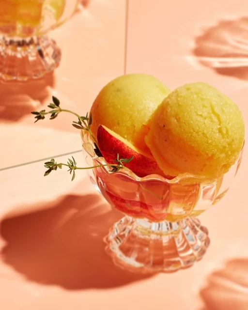 Close-up of yellow sorbet scoops served in a clear glass dessert cup with peach slice and thyme sprigs, against a peach-colored background.