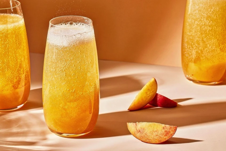 Sparkling beverage in a tall glass with fresh peach slices on a light surface, featuring bright lighting and an orange background.