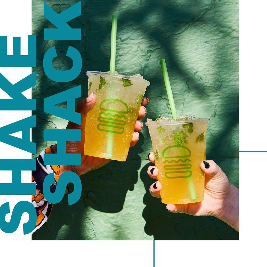 Two hands hold up clear plastic cups with orange liquid, green straws, and mint garnish, set against a light green backdrop.