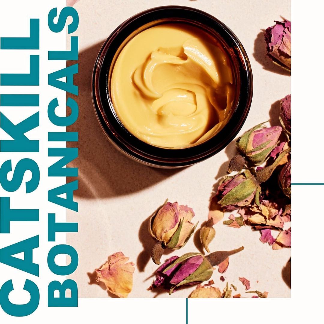 Overhead shot of a skincare product in a brown glass jar surrounded by dried rosebuds against a pale background, close-up, studio lighting.
