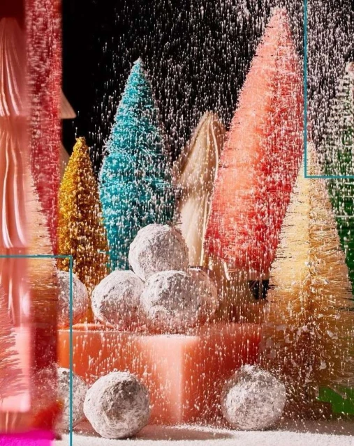 Colorful bottle brush trees covered in faux snow and powdered donuts sit on a pink platform in a Christmas holiday still life.