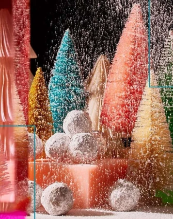 Colorful bottle brush trees covered in faux snow and powdered donuts sit on a pink platform in a Christmas holiday still life.