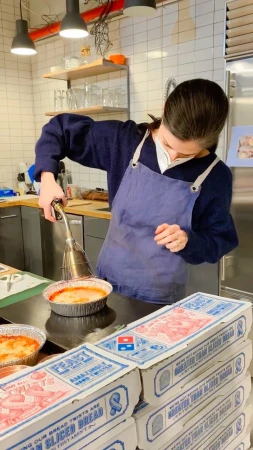 Woman in apron using a torch on a food item, surrounded by Domino's bread boxes, indoors under warm lighting, in a food preparation area.