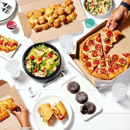 Overhead shot of Domino's pizza, pasta, salad, sandwiches, bread bites, and lava cakes arranged on a white wooden table, ready to eat.