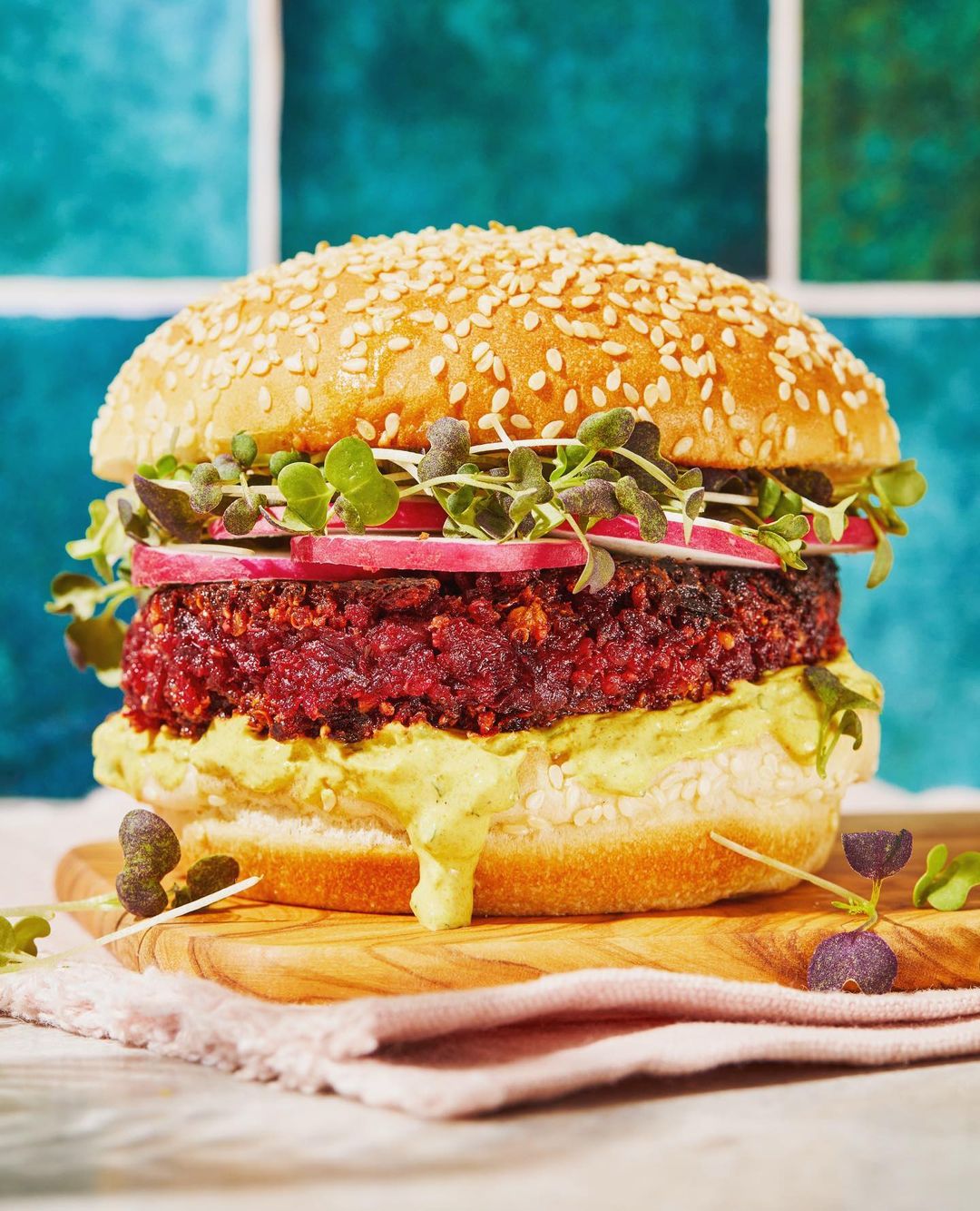 Veggie burger on a wooden board, topped with sprouts and sliced radishes, against a teal tile backdrop, showcasing a healthy food option.