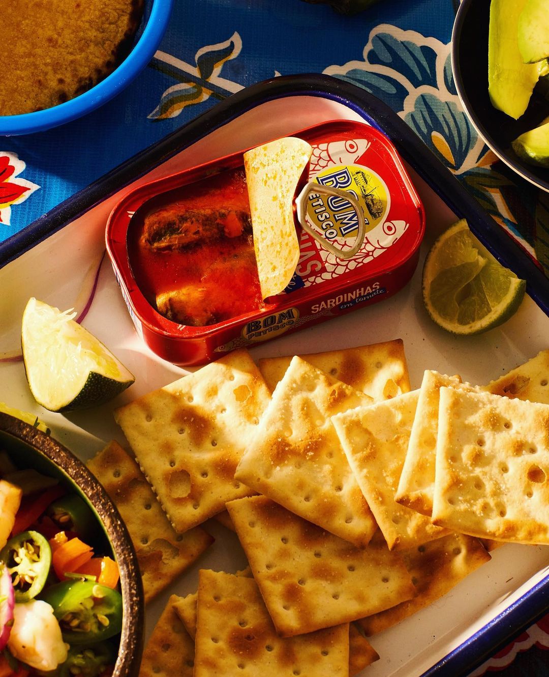 An open can of sardinhas served on a white tray with crackers and lime slices. Food and beverage photography featuring a colorful arrangement.
