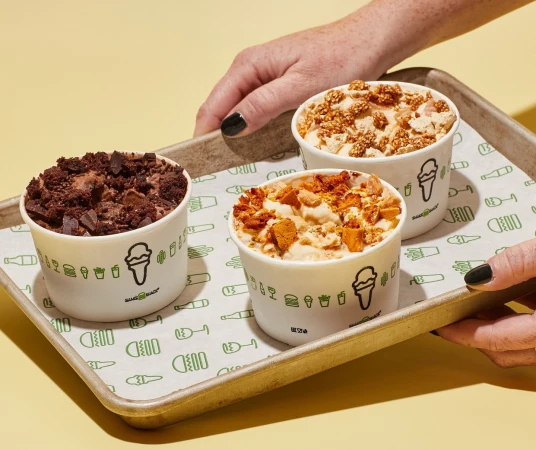 Three paper cups filled with assorted ice cream sit on a metallic tray held by a person's hand against a yellow backdrop.