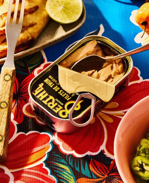 Overhead food shot of an opened can of tuna with a spoon, lime slice, grilled flatbread, and guacamole on a floral tablecloth.