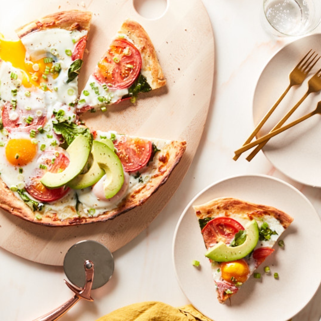 Overhead shot of a breakfast pizza with sliced avocado, tomatoes, green onions, spinach, and eggs on a wooden cutting board.