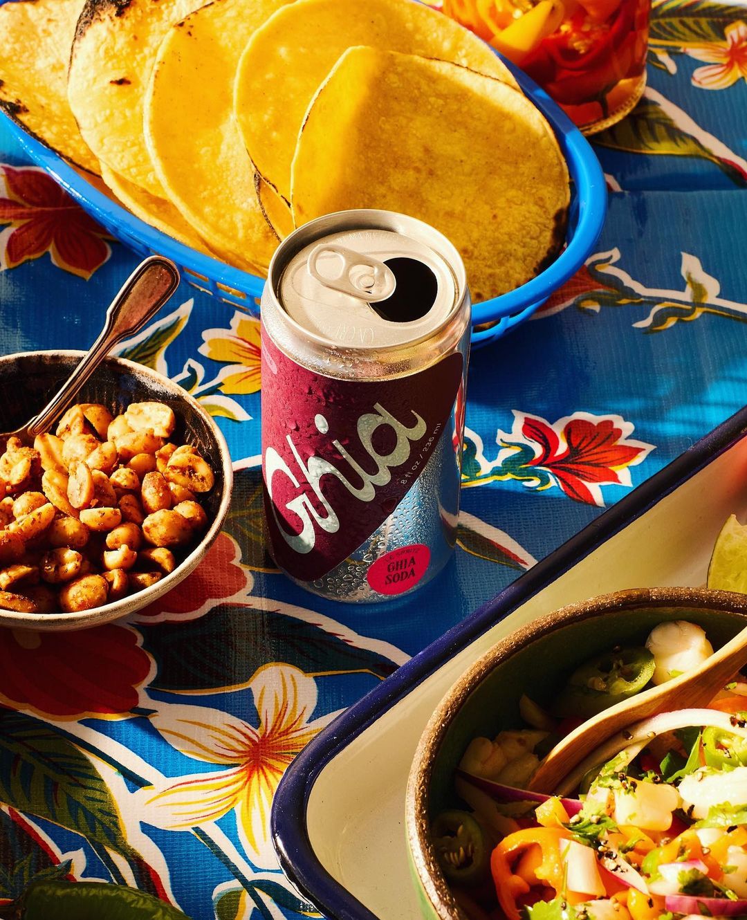A can of Ghia soda sits amidst a colorful table setting with tortillas, peanuts, and a taco, bathed in bright, natural light.