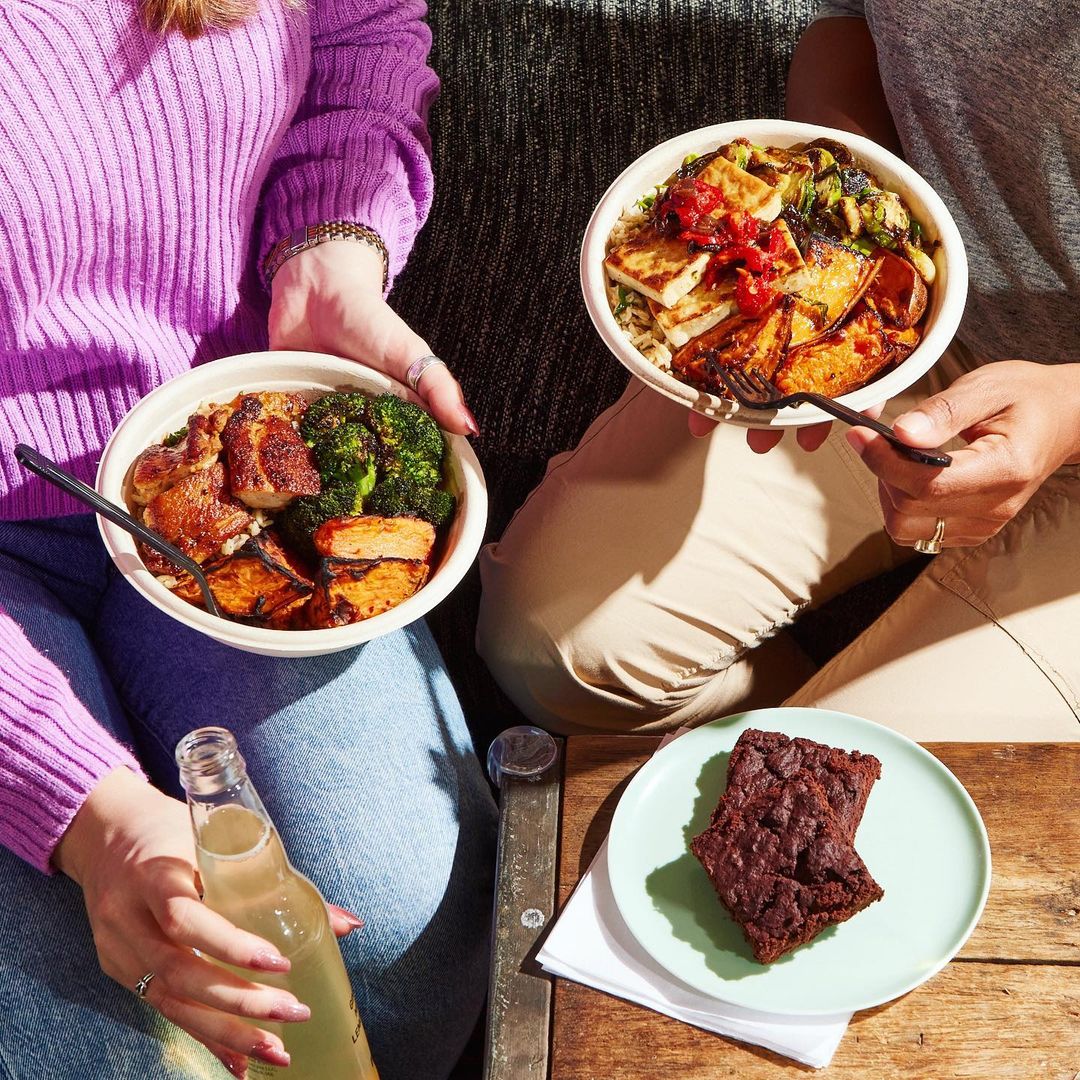 Two people sitting together and holding bowls of prepared food, with a brownie on a plate and a bottle of sparkling lemonade.