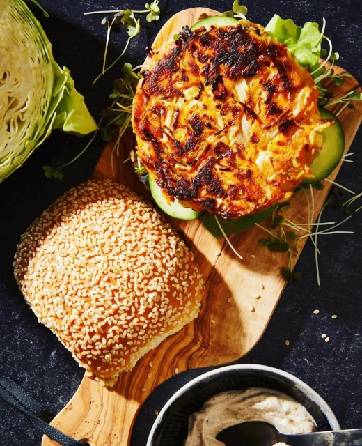 Overhead shot of a plant-based burger with a sesame seed bun, cabbage, sauce and sprouts served on a wooden cutting board.