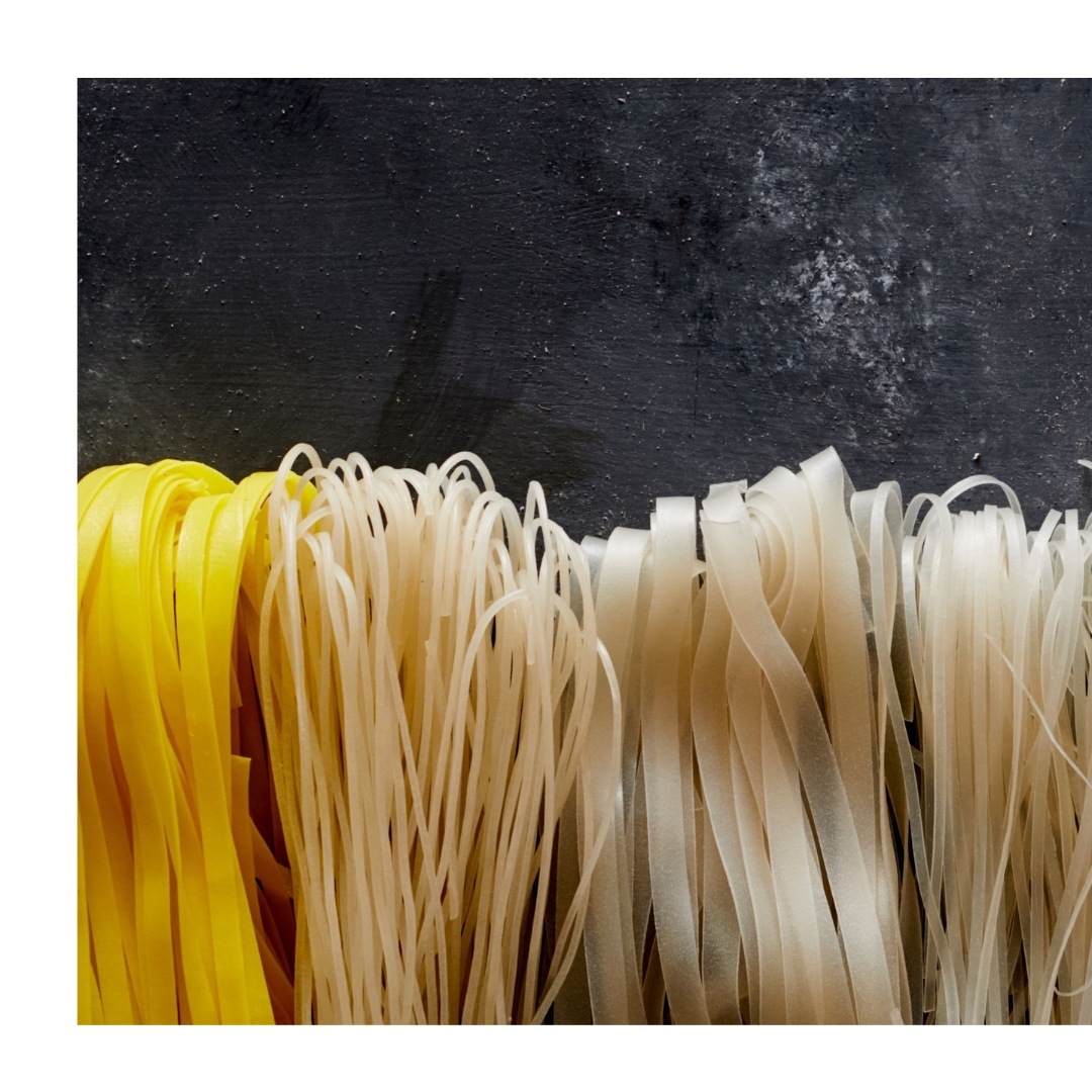 High-angle shot of assorted uncooked noodles: yellow fettuccine, cream-colored round noodles, and white rice noodles against a dark backdrop.
