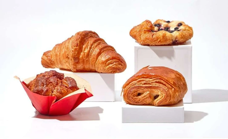 Close-up of croissant, muffin, and pastries on white cubes and a bright white surface, arranged in a food still life photograph.