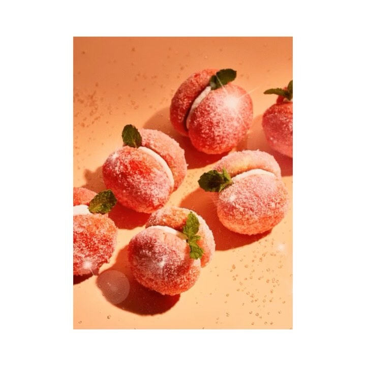 Peach-shaped pastries with cream filling and mint leaves, dusted with sugar, are displayed on a light orange background.