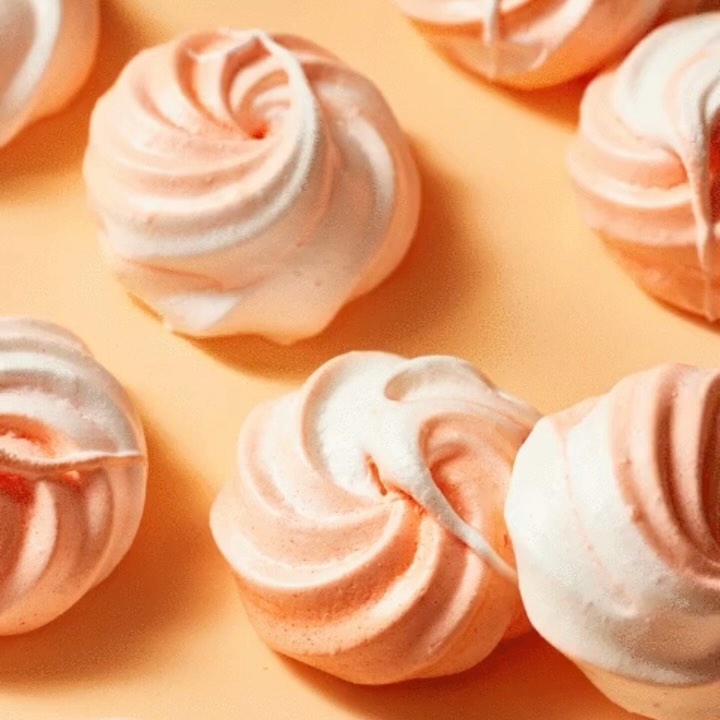 Close-up of pink and white meringues arranged on a peach-colored surface, highlighting their delicate swirl pattern and pastel hues.