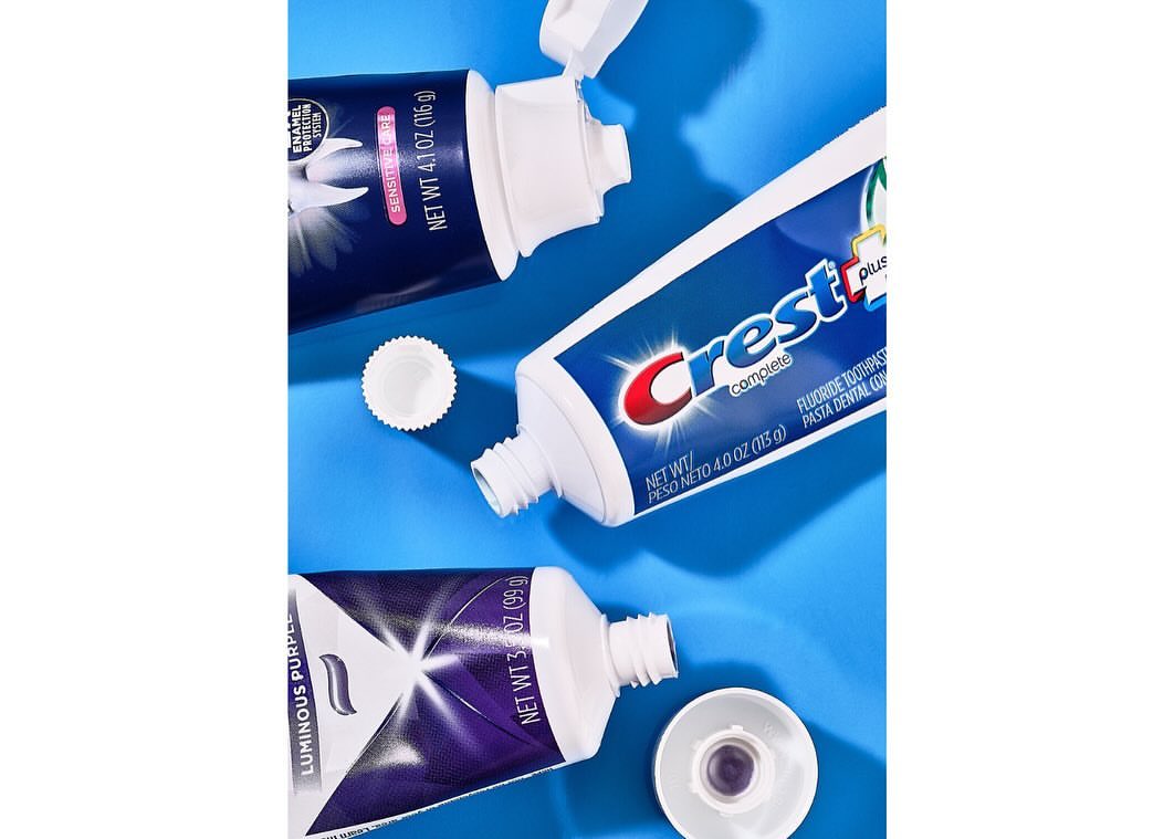 Three tubes of toothpaste, including Crest Complete and Luminous Purple varieties, arranged on a light blue background with product information.