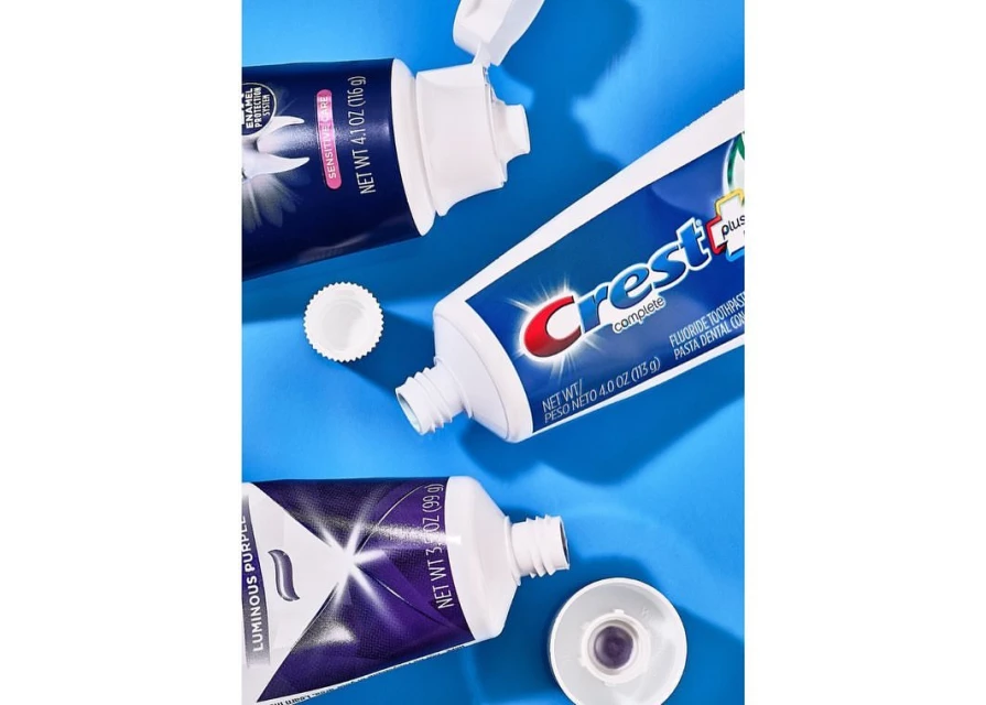 Three tubes of toothpaste, including Crest Complete and Luminous Purple varieties, arranged on a light blue background with product information.
