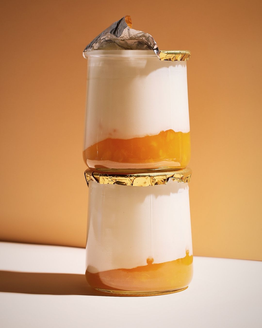 Two stacked glass jars filled with layers of white yogurt and orange fruit compote against a warm orange background in a studio setting.