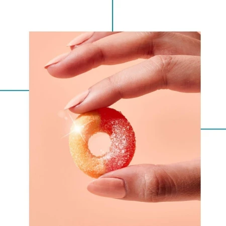 A brightly lit, close-up studio shot of a human hand holding a frosted peach ring candy against a peach-colored background.