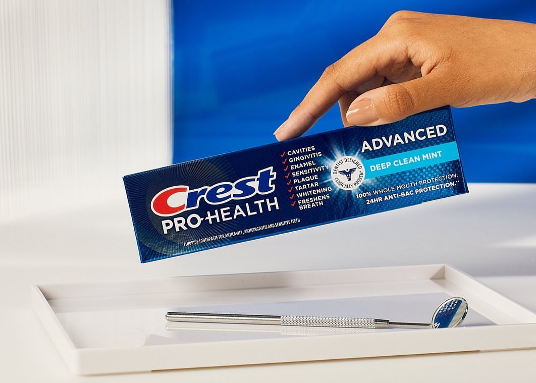 Crest Pro-Health Advanced Deep Clean Mint toothpaste box held by a hand on a white tray with a dental mirror, set against a blue backdrop.