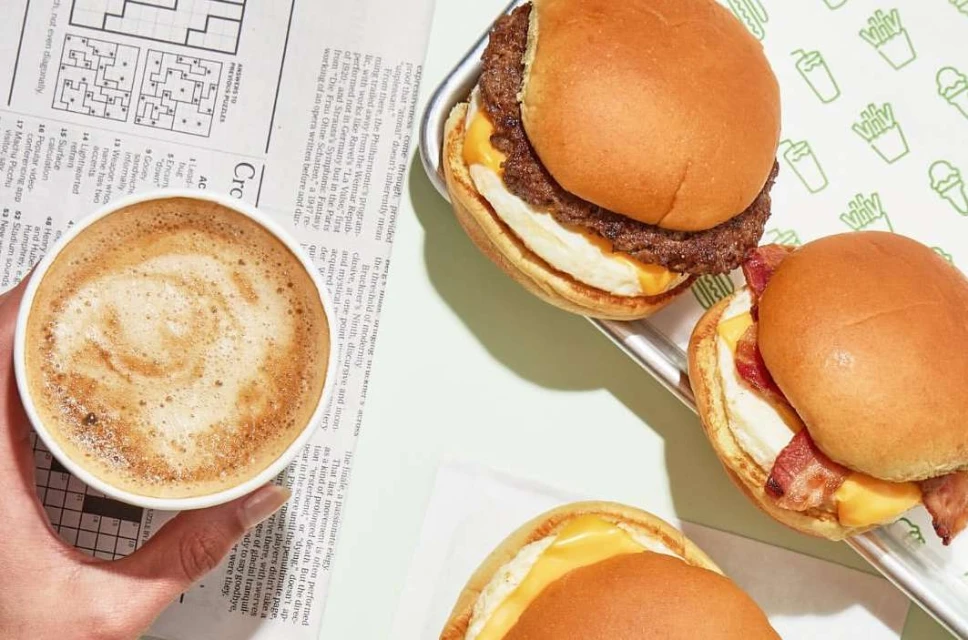 Overhead shot of three breakfast sandwiches on a tray, with a coffee cup on a newspaper, and the Shake Shack logo in the background.
