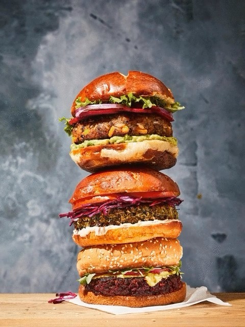 Stack of three vegetarian burgers with various toppings and sesame buns on a wooden surface against a dark grey background.