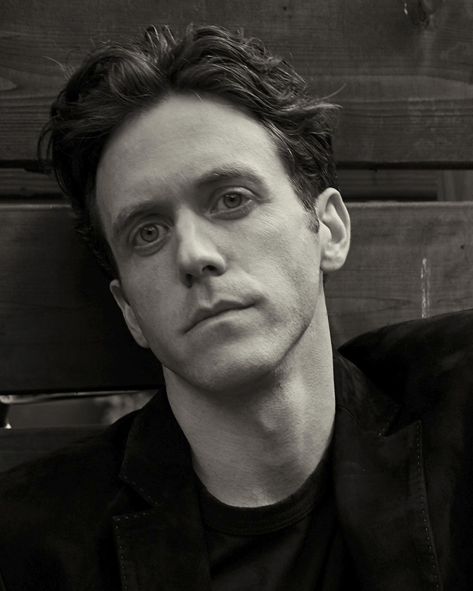 Headshot of an adult man with dark hair wearing a blazer against a weathered wood background in black and white with eye-level angle.